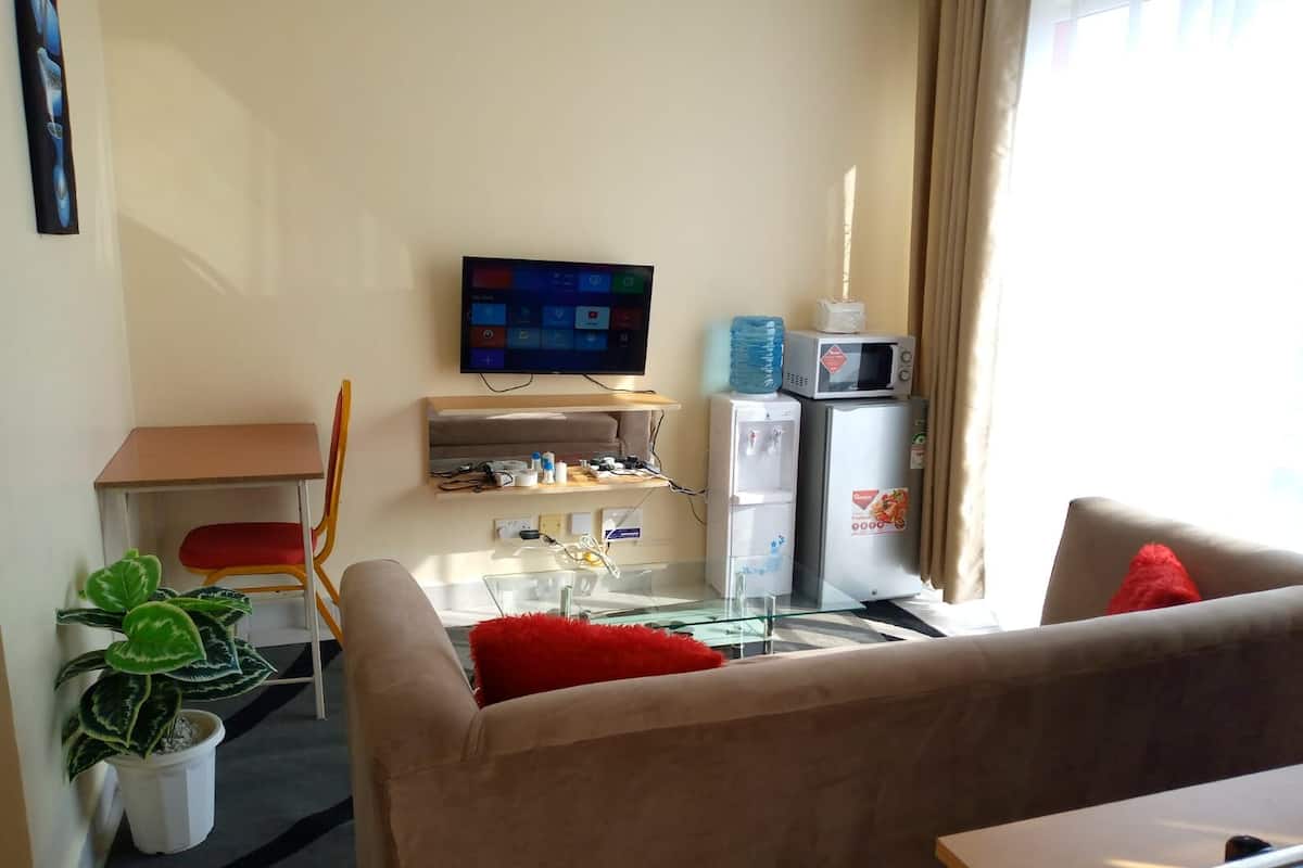 A living room with a flat-screen TV, a water dispenser, a microwave, a sofa, a small table, and a potted plant.