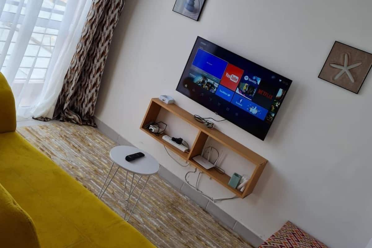 A living room with a yellow sofa, a TV mounted on the wall displaying various streaming services, a small table with a remote control, and a patterned rug.
