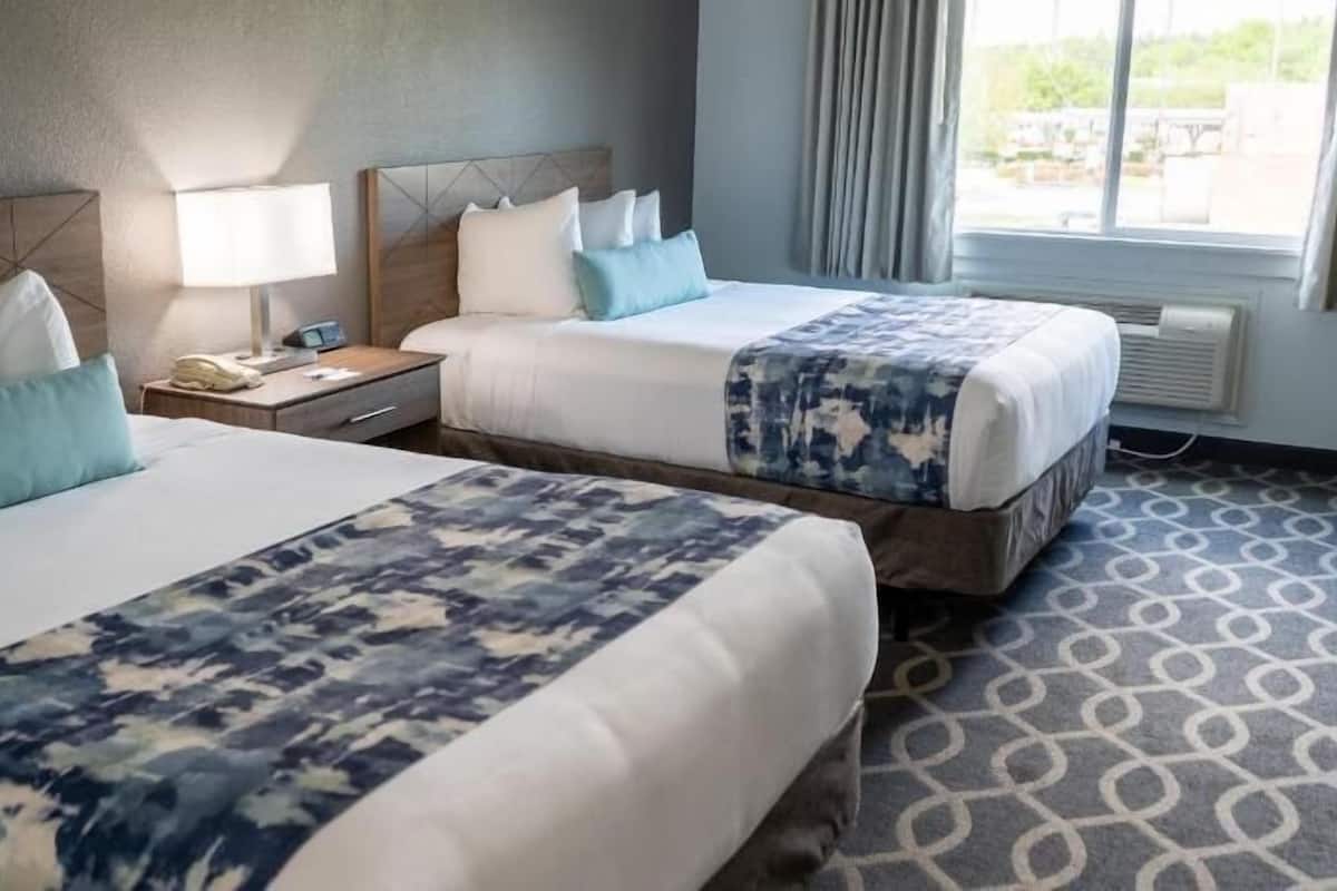 A hotel room with two beds, a nightstand, a lamp, and a window with curtains.