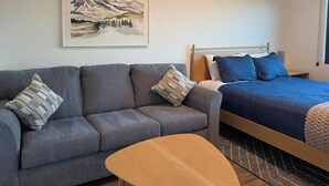 Et moderne værelse med en grå sofa, et træbord, en seng med blåt sengetøj og et maleri af et bjerglandskab på væggen.