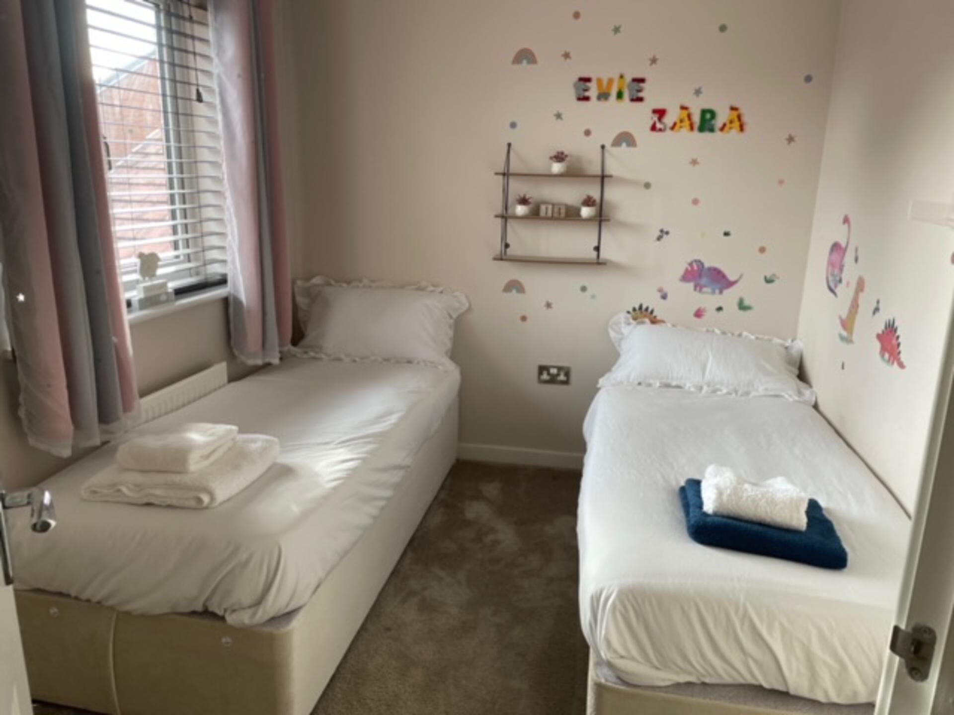 Ein Schlafzimmer mit zwei Betten, einem fenster mit Vorhängen, einem Regal mit Dekoration und Wandbildern.