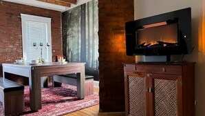 A cozy room with a brick wall, a fireplace, a TV mounted on a wooden cabinet, and a dining table with benches. - Historic Haven Hideaway California King Bed

 (Cleveland)