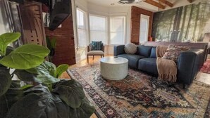 A living room with a blue sofa, a round coffee table, and a patterned rug. There are large windows with blinds and a brick wall. - Historic Haven Hideaway California King Bed

 (Cleveland)
