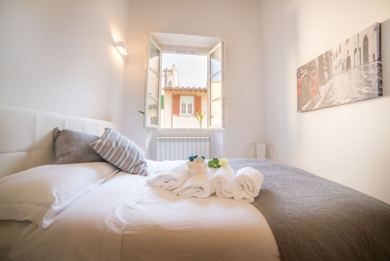 A hotel room with a bed, a window with a view of buildings, a radiator, and a wall-mounted picture.
