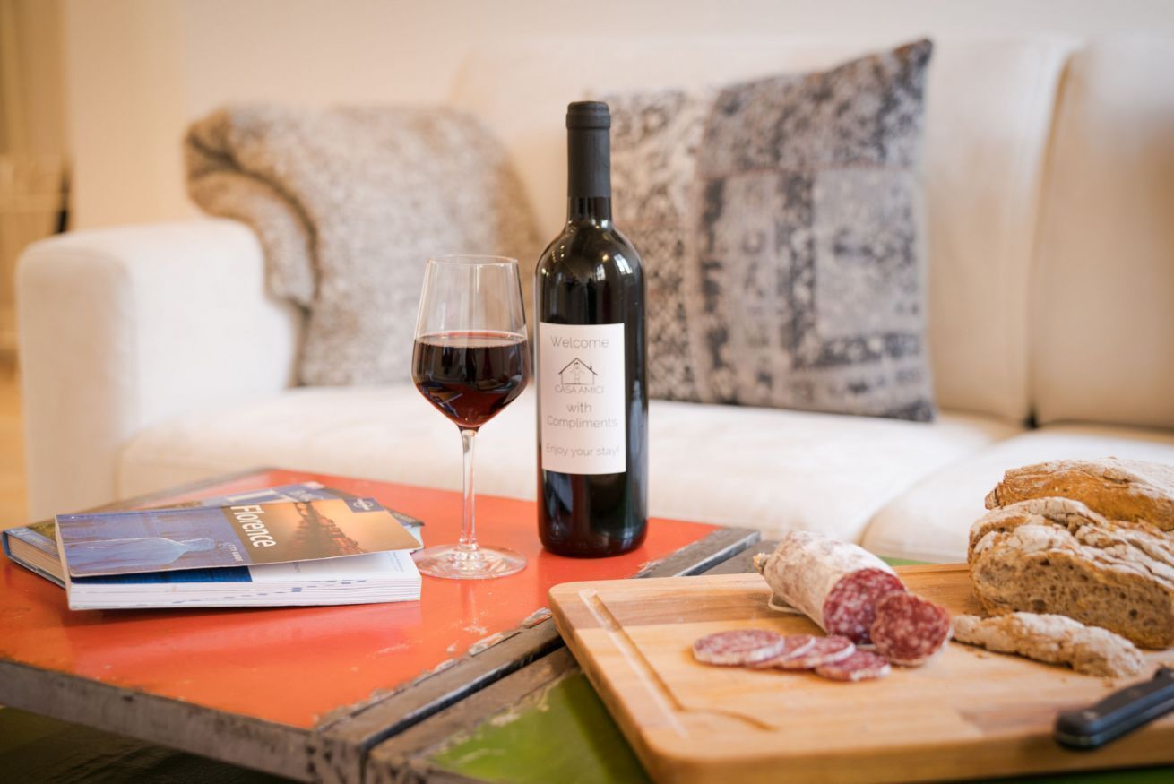 A bottle of wine, a glass of red wine, a book, a cutting board with sliced salami, and a loaf of bread on a table.