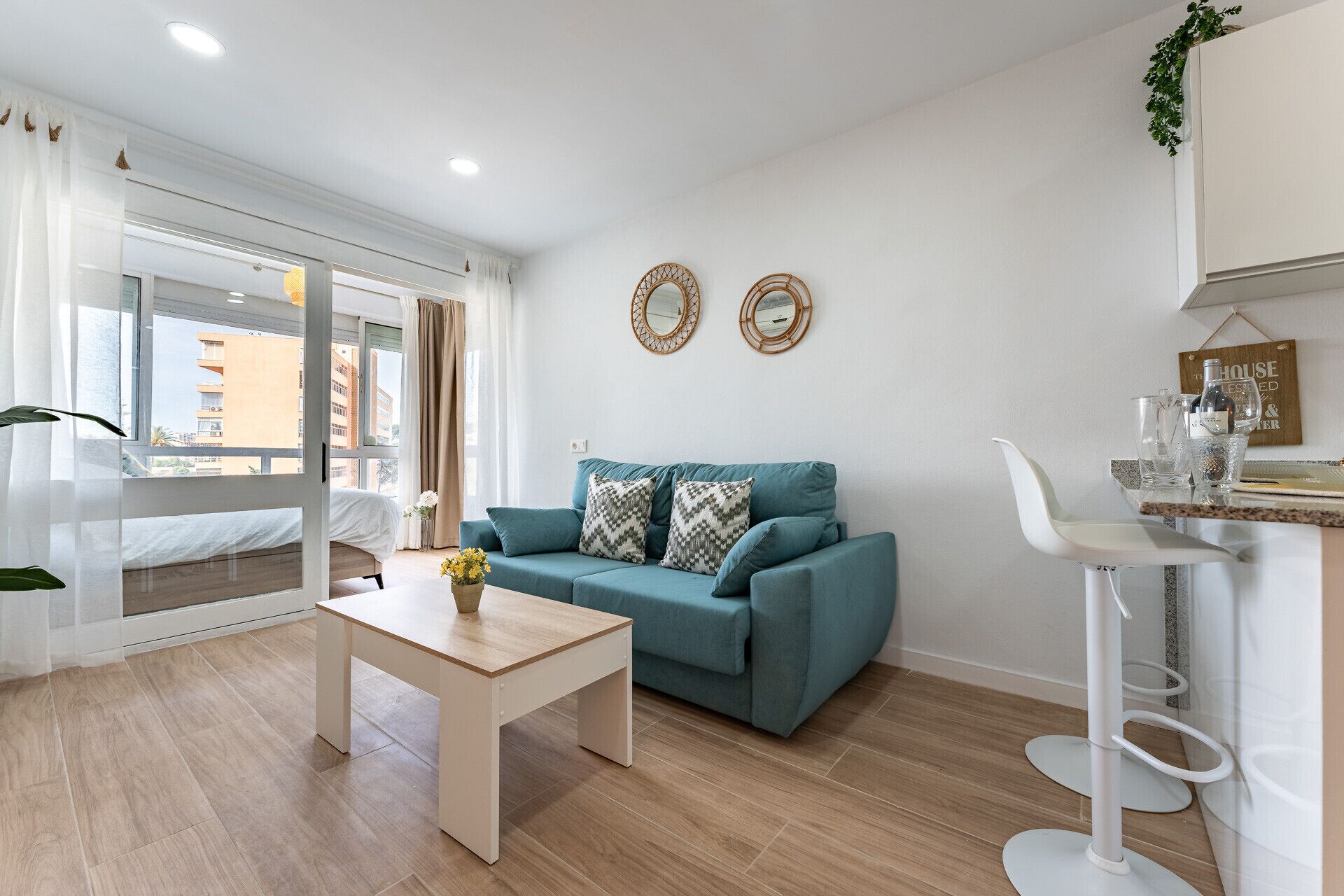 Una sala moderna con un sofá color turquesa, una mesa de centro de madera y una silla blanca. Hay dos espejos decorativos en la pared y una planta pequeña en el alféizar de la ventana.