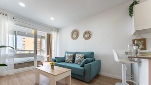 A modern living room with a teal sofa, a wooden coffee table, and a white chair. There are two decorative mirrors on the wall and a small plant on the windowsill. - MalagaSuite Cozy Studio & Pool Torremolinos (Torremolinos)