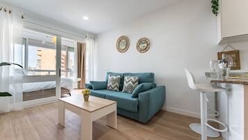 Una sala moderna con un sofá color turquesa, una mesa de centro de madera y una silla blanca. Hay dos espejos decorativos en la pared y una planta pequeña en el alféizar de la ventana.