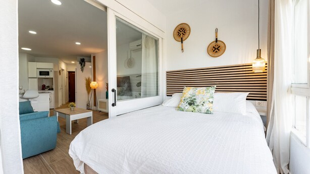 A modern bedroom with a large bed, a glass door leading to a balcony, a small table, and a view of the kitchen. - MalagaSuite Cozy Studio & Pool Torremolinos (Torremolinos)