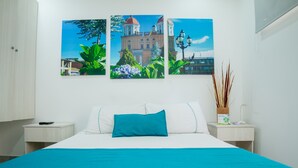 A hotel room with a bed, two bedside tables, a nightstand, a lamp, and three wall-mounted paintings. - Almaviva Casa Hotel (Medellín)