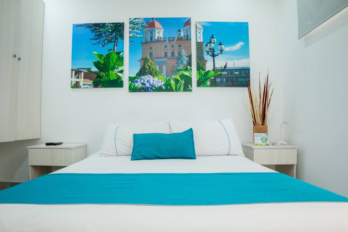 Une chambre d’hôtel comprenant un lit, deux tables de chevet, une petite table, une lampe et trois tableaux accrochés au mur.