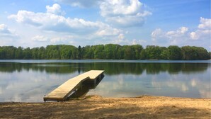 Une jetée en bois s’étendant dans une eau calme, bordée d’une plage de sable et d’une rive boisée.
