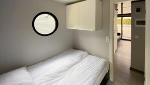 Une petite chambre avec un lit simple, une fenêtre ronde, une porte blanche donnant sur un balcon et un interrupteur sur le mur.