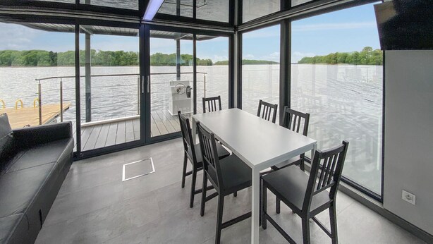 Un espace de restauration moderne avec une table et des chaises blanches, de grandes fenêtres offrant une vue sur un lac paisible et une végétation luxuriante.