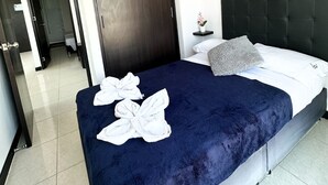 Une chambre d’hôtel avec une porte en bois foncé, un lit doté d’une tête de lit noire, du linge de lit blanc, une couverture bleue et des serviettes disposées sur le lit.