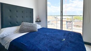 Une chambre à coucher avec un grand lit, une table de chevet et une vue sur un balcon d’où l’on aperçoit une tour au loin.