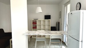 Eine Küche mit einem weißen Kühlschrank, einem Esstisch mit zwei Stühlen, einer weißen Pendelleuchte und einem Fernseher auf einem Regal.