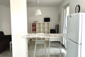 A kitchen with a white refrigerator, a dining table with two chairs, a white pendant light, and a television on a shelf.