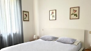 Ein ordentlich bezogenes Bett mit Streifenmuster, eine Nachttischlampe und zwei gerahmte Pflanzenbilder an der Wand.