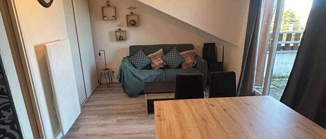 A cozy living area with a sofa, a dining table, and chairs. There is a kitchen area with a stove and cabinets. The room has a sloped ceiling with hanging lights.