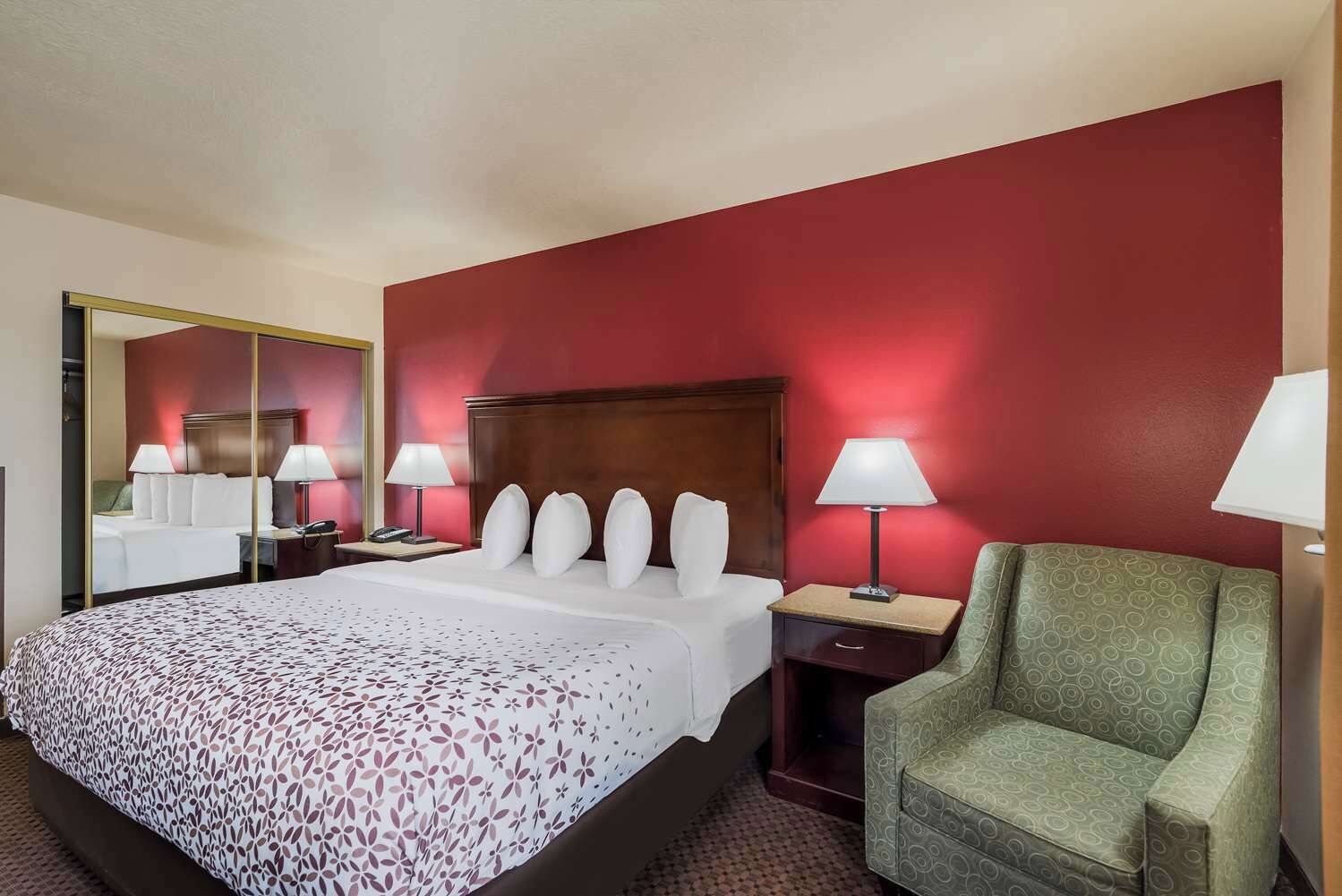 Une chambre d’hôtel avec un grand lit, deux tables de chevet avec des lampes, un fauteuil vert et un miroir au mur.