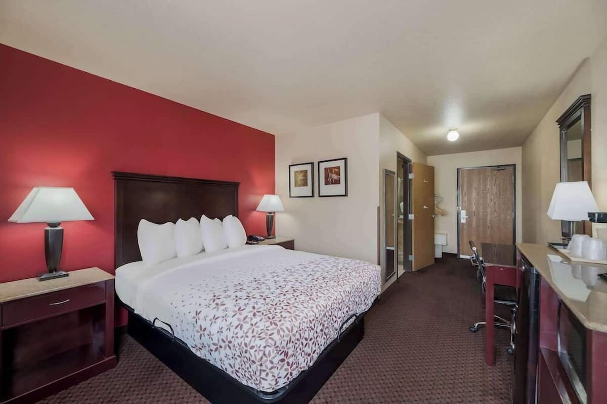 A hotel room with a large bed, two bedside tables with lamps, a desk with a chair, a mirror, and a door leading to another room.
