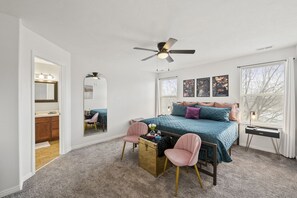 A bedroom with a bed, two chairs, a desk, a ceiling fan, and a window with blinds.
