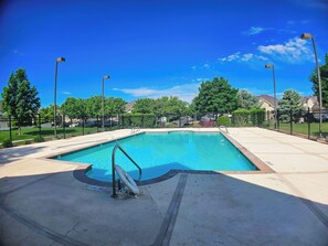 A rectangular swimming pool with a diving board and a lifeguard chair, surrounded by a fence, trees, and houses in the background.