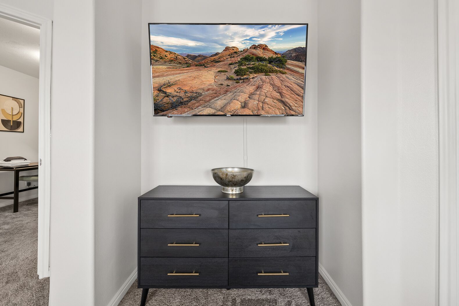 Een gang met een donker houten kastje, een schaal erbovenop en een lijst met een landschapsfoto aan de muur.