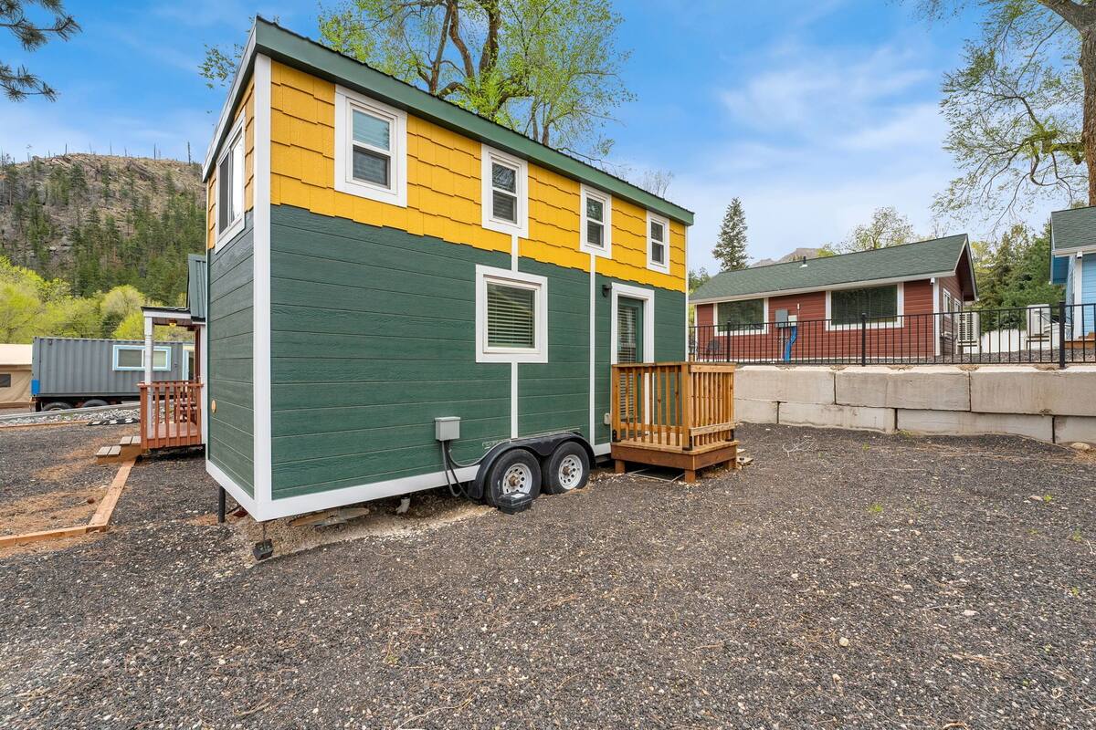 A small, two-story mobile home with a green and yellow exterior, a wooden deck, and a trailer attached.