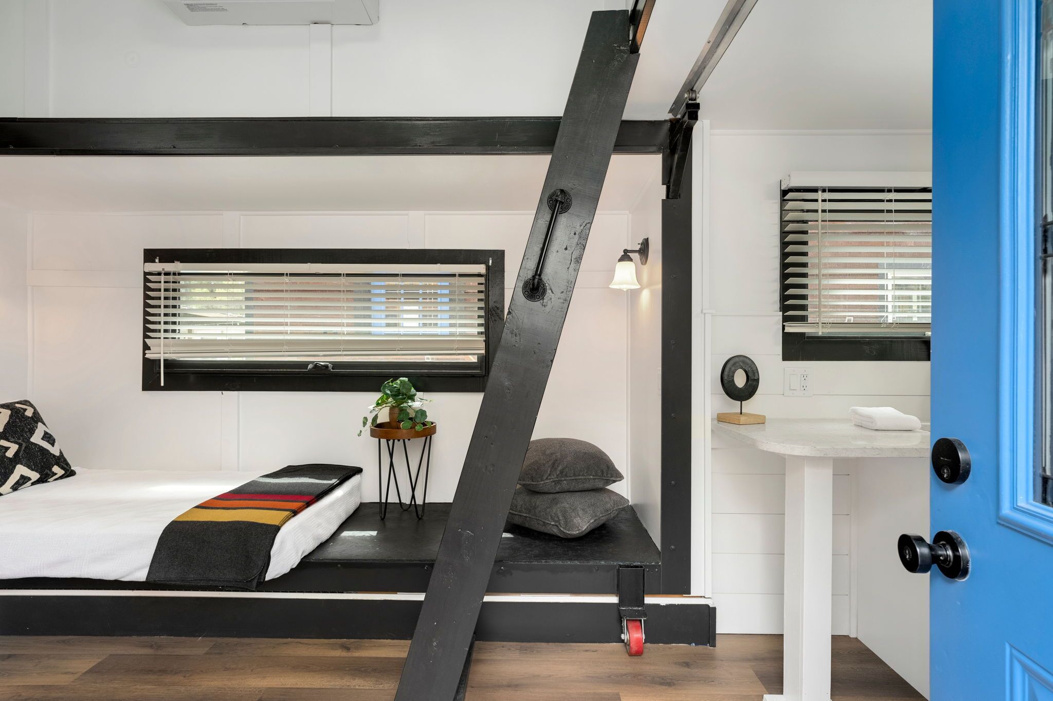 Ein kleines Zimmer mit einem Bett, einem kleinen Nachttisch mit Pflanze, einem Fenster mit Jalousien, einem weißen Waschbecken und einer blauen Tür.