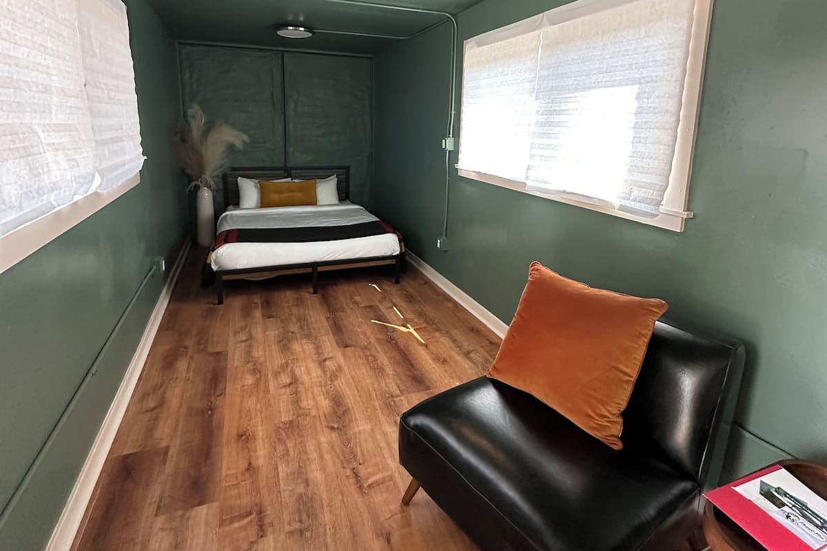 A room with a bed, a black leather chair, a wooden table, and a window with blinds.