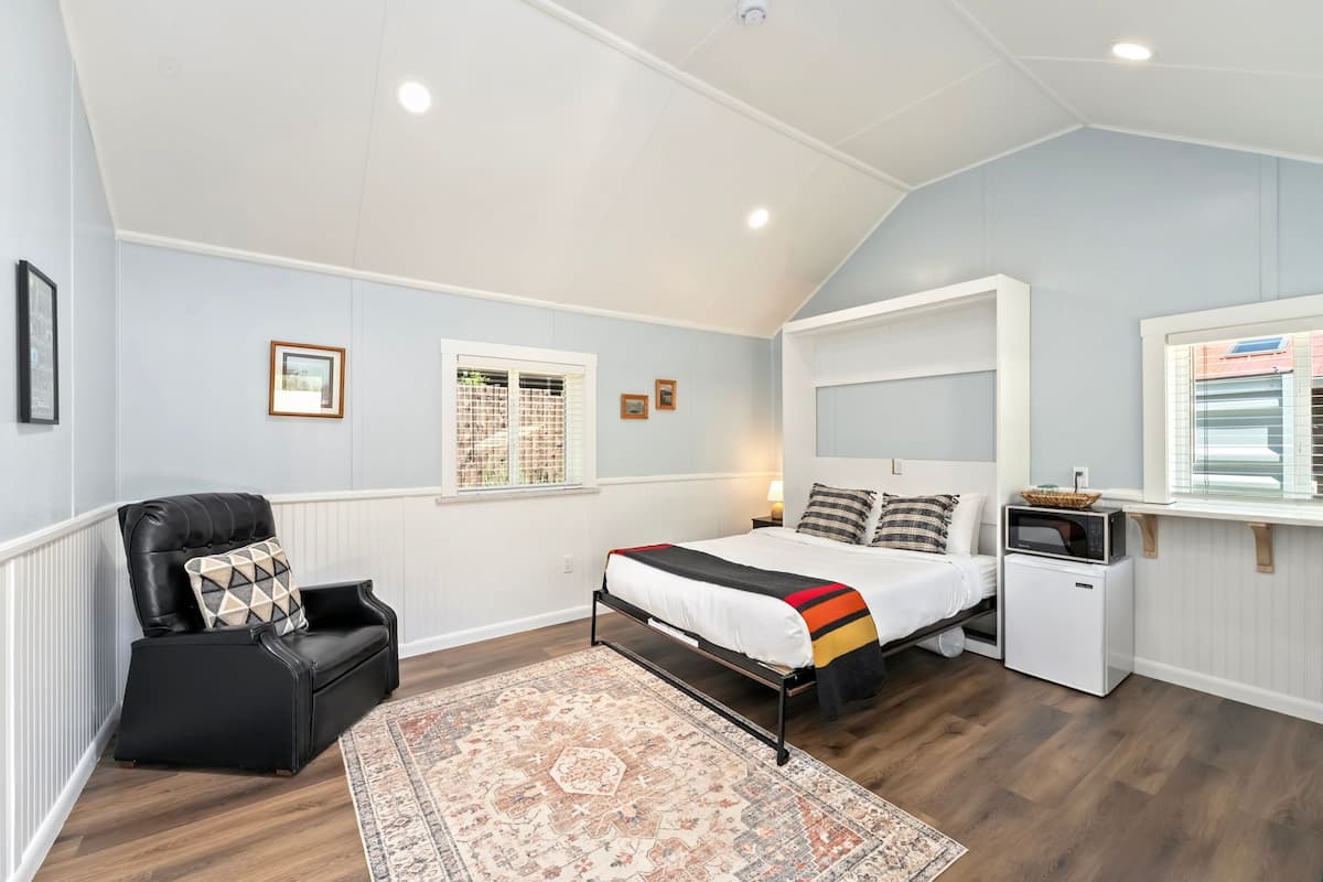 A bedroom with a bed, a black leather chair, a small white appliance, and a window with blinds.
