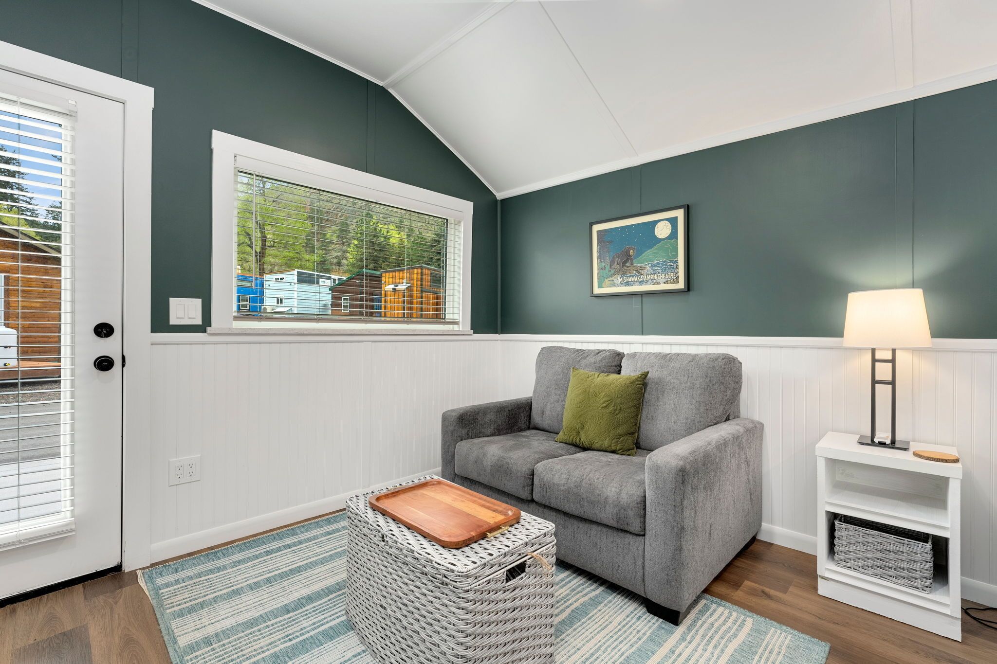 A room with a grey sofa, a wooden tray, a wicker basket, a lamp, and a window with blinds.
