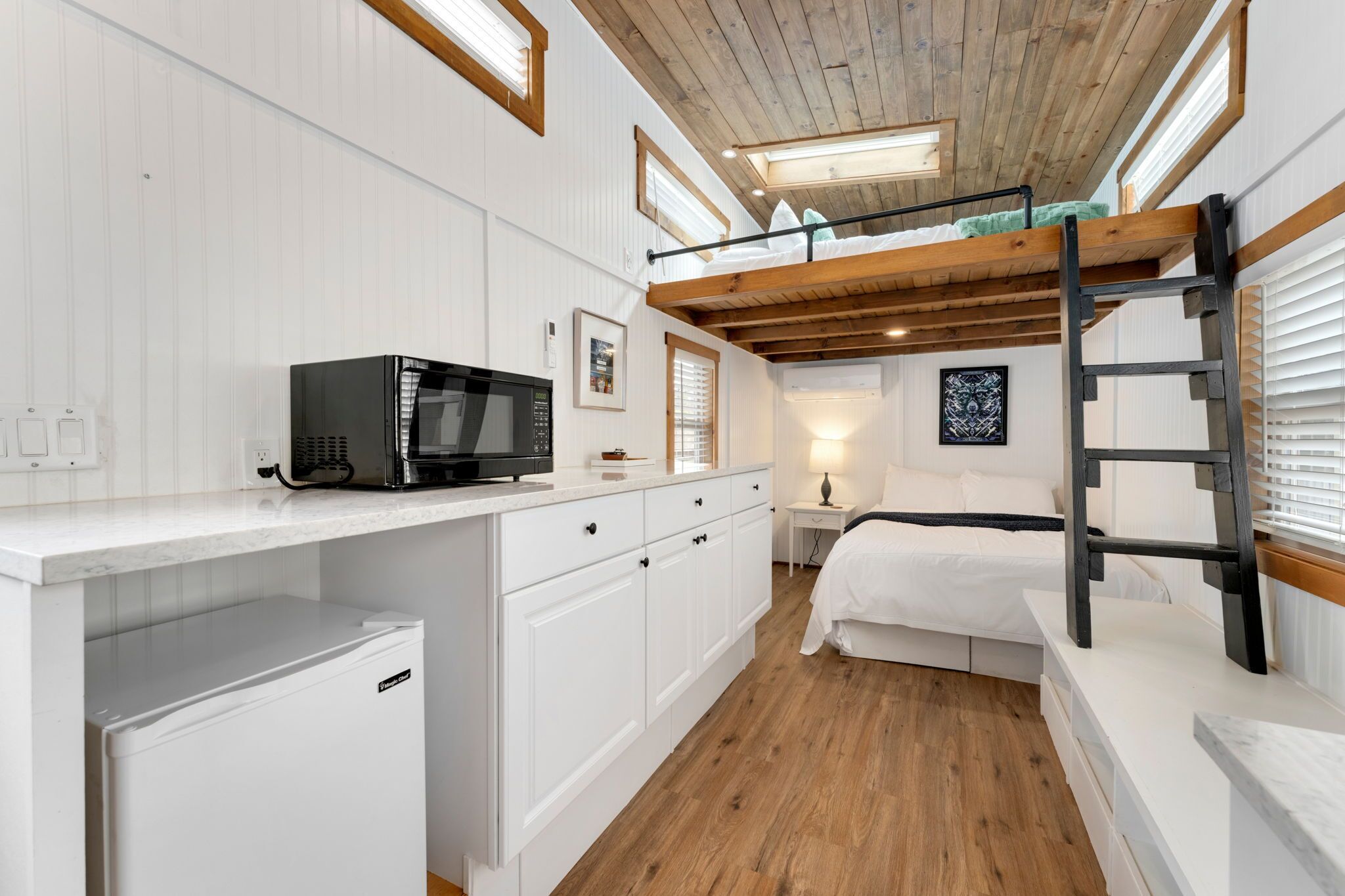 Habitación pequeña con literas, una kitchenette equipada con microondas y un refrigerador compacto, y una mesita de noche con lámpara.