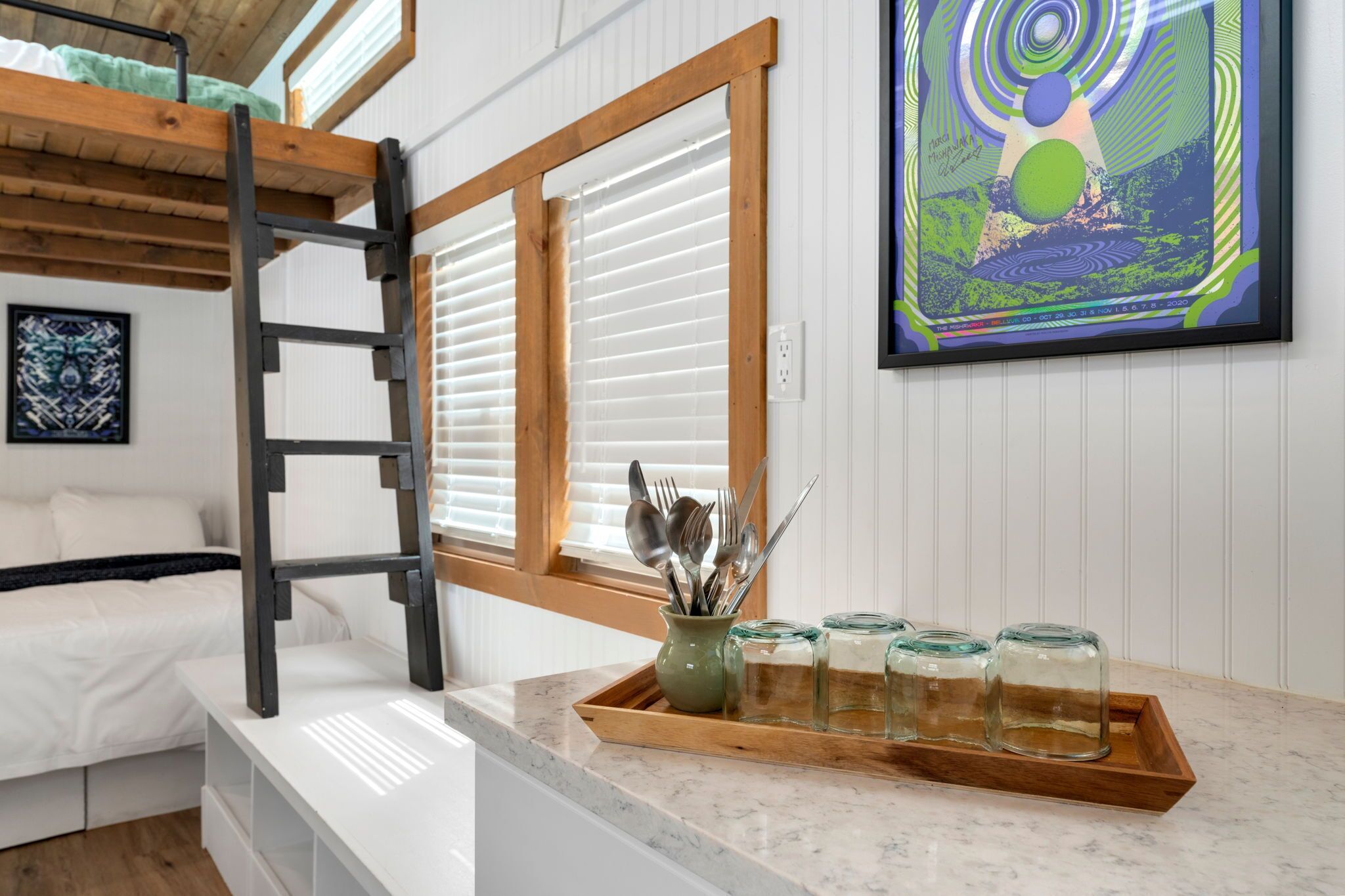 A bunk bed with a ladder, a window with blinds, a tray with jars, and a framed artwork on the wall.