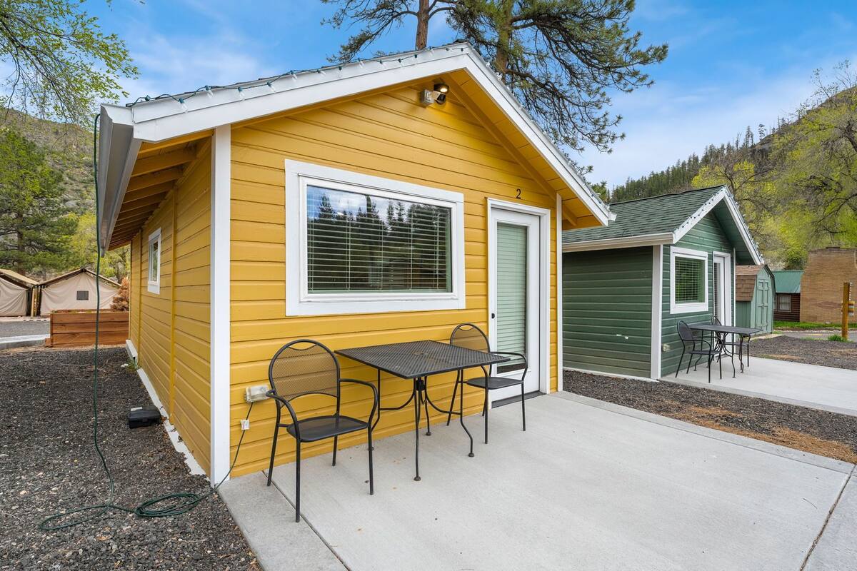 A yellow cabin with a patio table and chairs, a green door, and a small window.