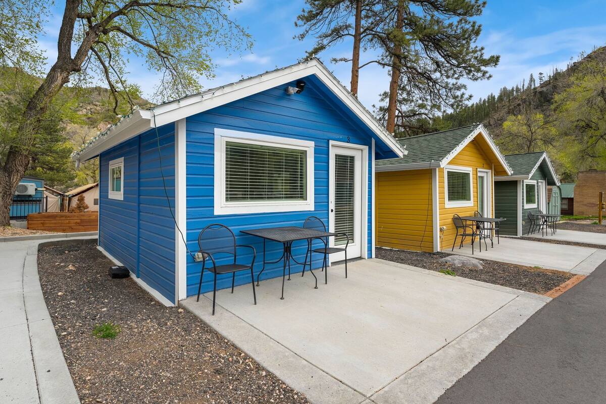 A blue cabin with a patio area, including a table and chairs, surrounded by trees and a hilly landscape.