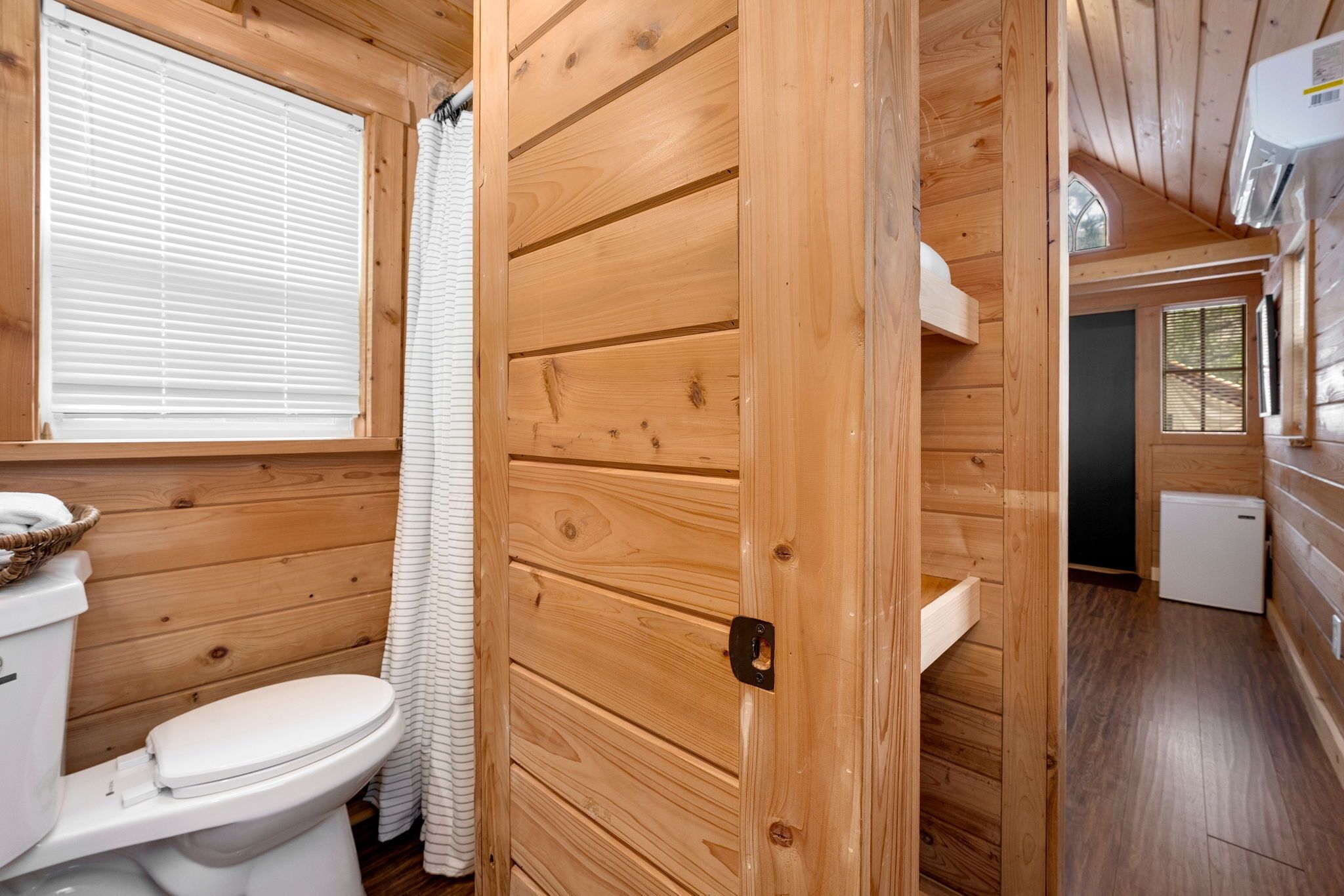 A wooden-paneled bathroom with a toilet, a sink, and a window with blinds.