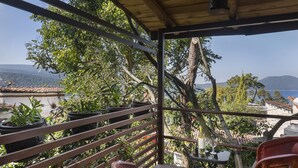 A balcony with potted plants, a table with a bottle and glass, and a view of the sea and hills.