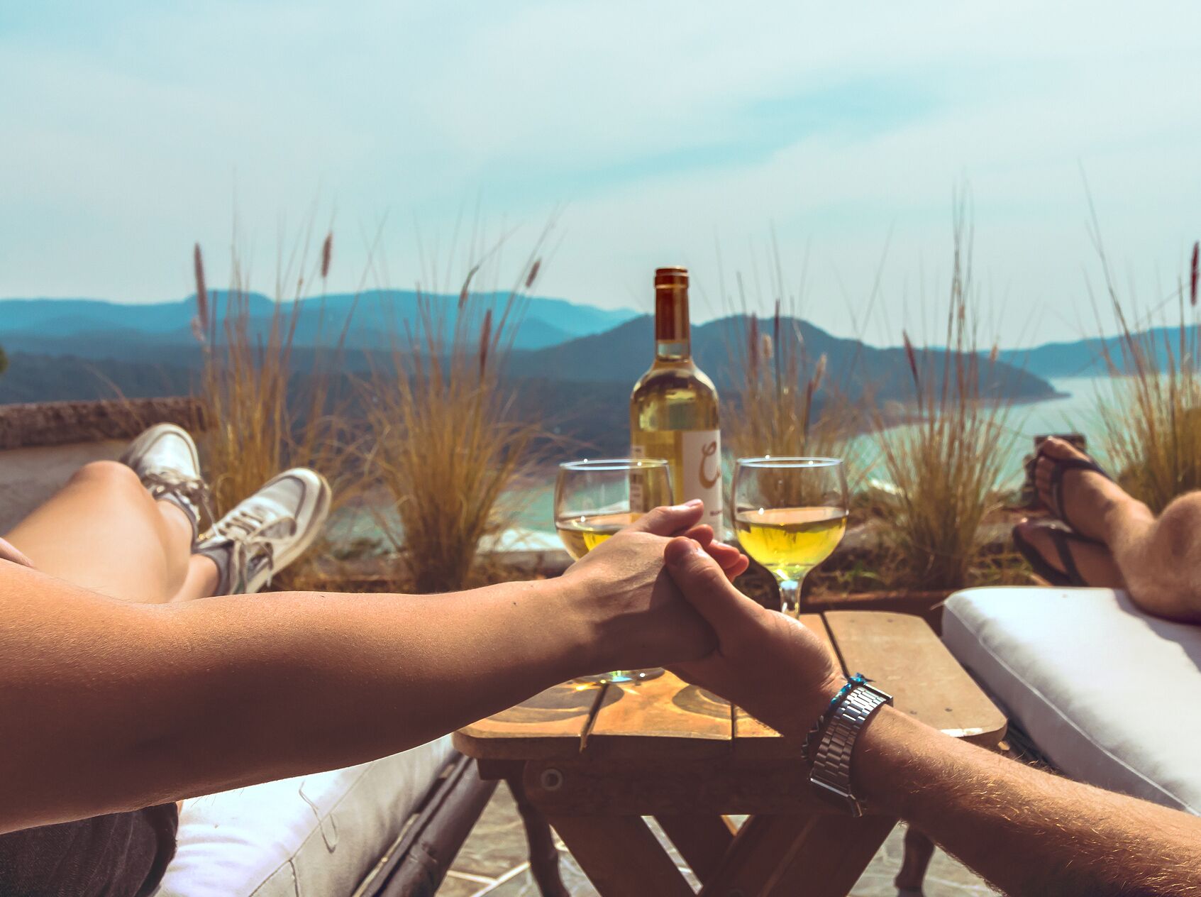 Two people holding hands at a table with a bottle of wine and glasses, overlooking a scenic view of mountains and water.