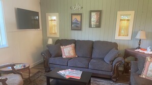 A living room with a dark grey sofa, a coffee table with magazines, a TV mounted on the wall, and framed pictures on the wall.