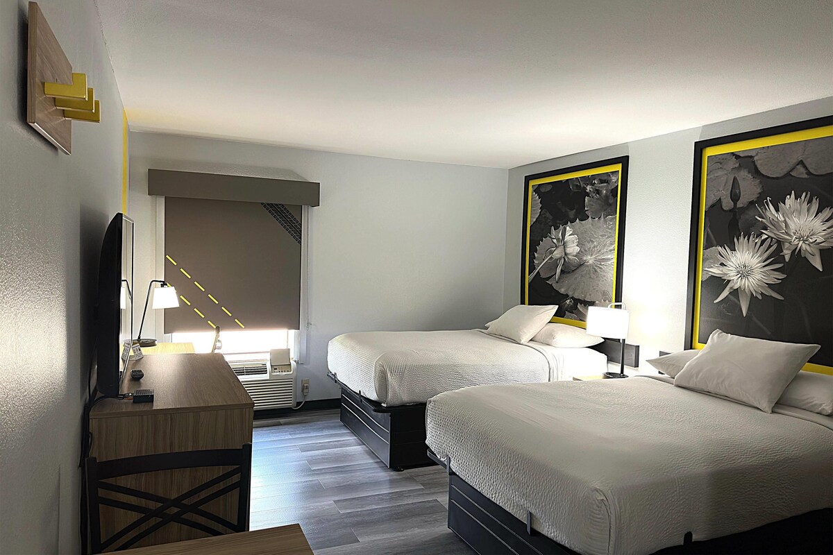 A hotel room with two beds, a desk, a chair, a TV, and artwork on the walls.