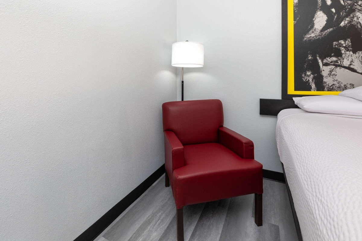 A hotel room with a red chair, a white bed, a floor lamp, and a framed artwork on the wall.