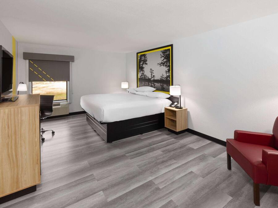 A hotel room with a bed, a desk with a flat-screen TV, a red chair, and a painting on the wall.