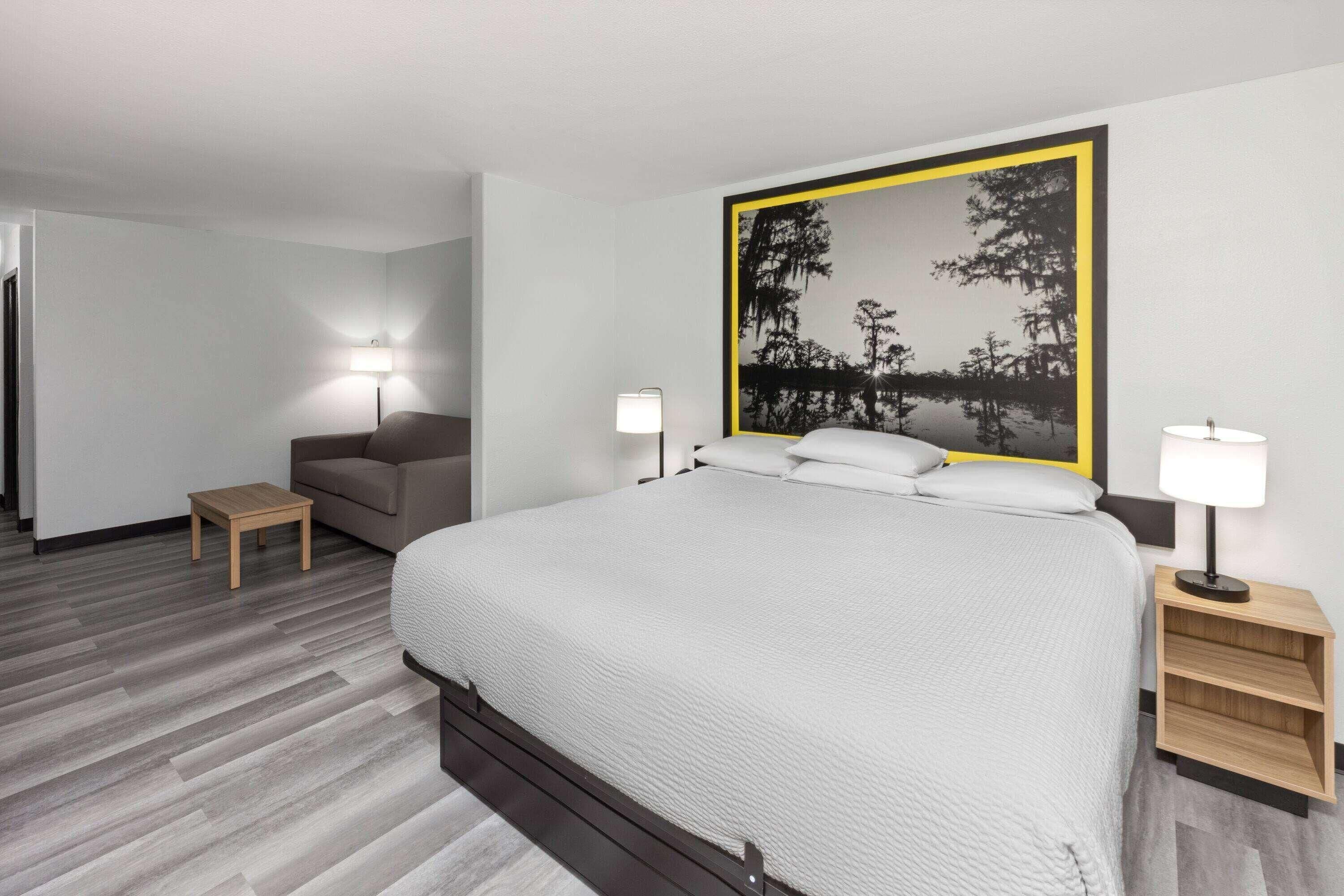 A modern hotel room with a large bed, two bedside tables with lamps, a small wooden side table, and a large framed picture on the wall.