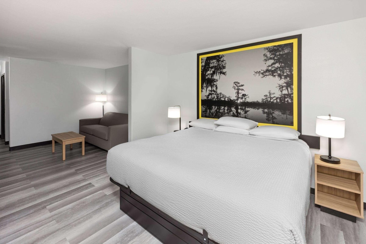 A modern hotel room with a large bed, two bedside tables with lamps, a small wooden side table, and a large framed picture on the wall.