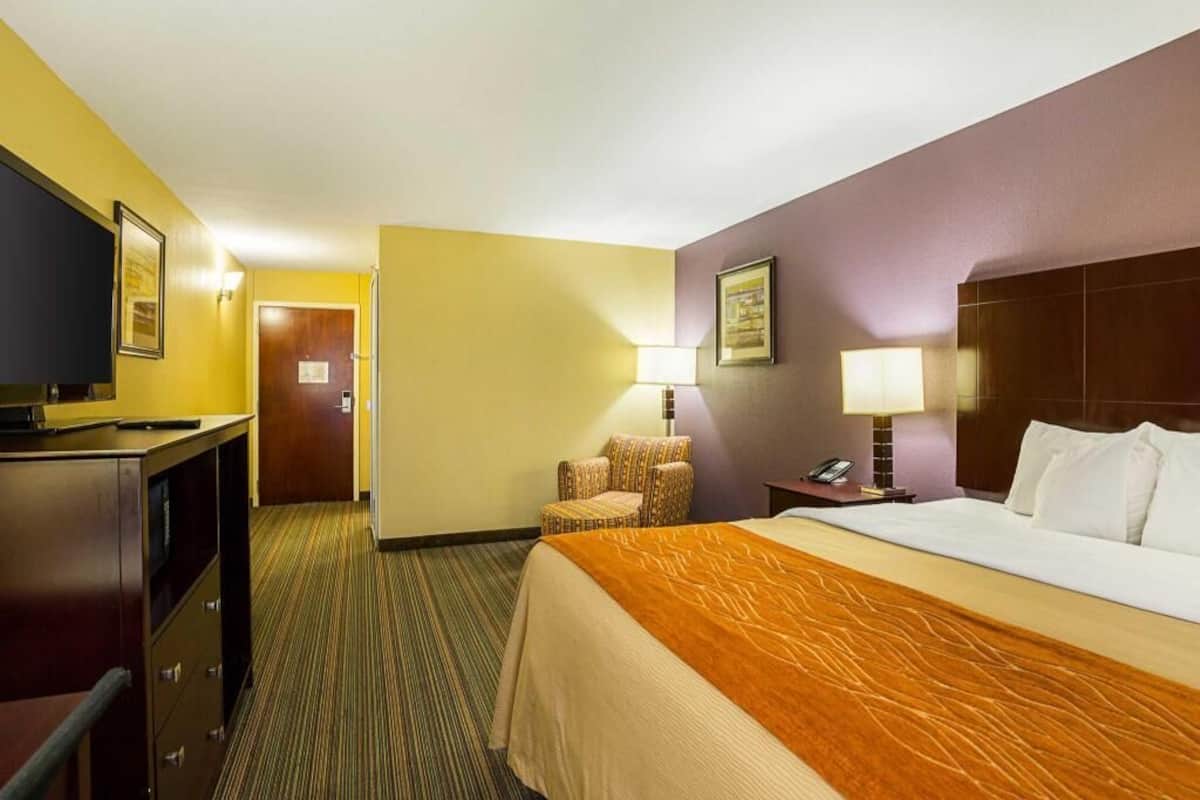 A hotel room with a bed, a desk, a chair, a lamp, and a television.