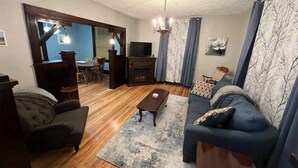 Een woonkamer met een bank, salontafel en open haard. Door een open doorgang is een eethoek met een tafel en stoelen zichtbaar.