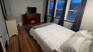 Een slaapkamer met een bed, een televisie op een houten standaard, een open haard en een raam met gordijnen.